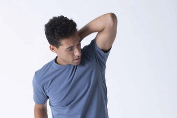Man checking his underarm sweat while wearing a blue T-shirt against a light background