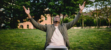 Man seated on a lawn with arms raised, wearing a light-colored undershirt and open jacket