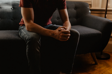 Person wearing a dark red T-shirt sitting on a couch with hands clasped