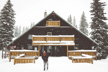 Person standing in winter clothing outside a snow-covered cabin during snowfall