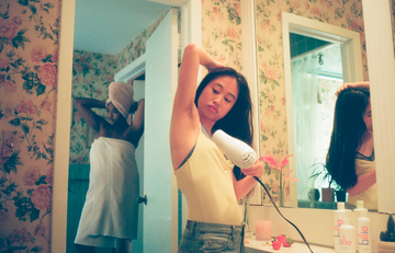 Woman drying her underarm with a hair dryer while another person towels off in a floral-wallpapered bathroom