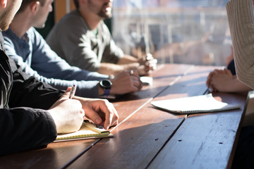 Colleagues take notes during a meeting around a sunlit wooden table