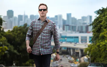 Man walking outdoors in a plaid shirt and sunglasses with a city skyline in the background