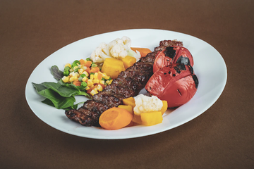 Angled view of a plate of steak with roasted vegetables, corn, potatoes, cauliflower, and red peppers on a brown table