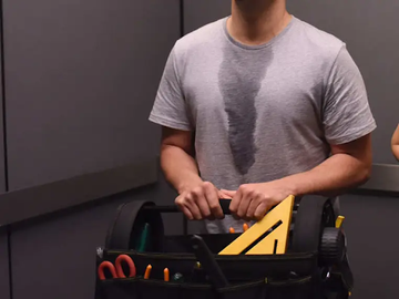 Person wearing a gray shirt with a large dark sweat stain holds a tool belt