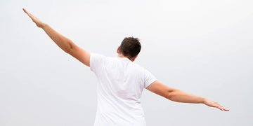 Man wearing a white undershirt with arms raised, viewed from behind against a light background