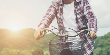 Man riding a bicycle in a white shirt and plaid overshirt with a basket on a sunny road